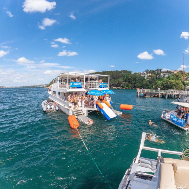 A group of people enjoy a sunny day on a large party boat with an inflatable slide into the blue water, surrounded by lush trees and a dock, with others swimming nearby.
