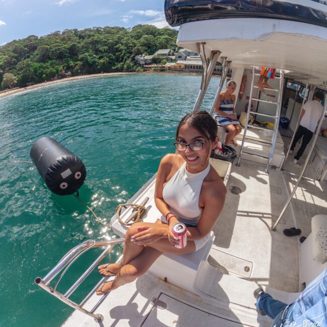 A woman in a white swimsuit sits smiling on a boat holding a can drink, with clear blue water and a forested shoreline in the background. Other people relax nearby under a sunny sky.