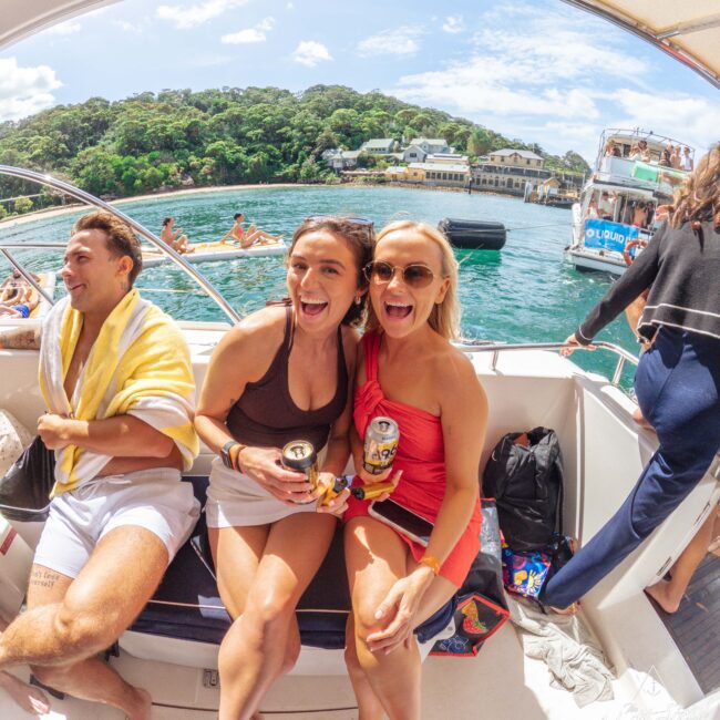 Two women in swimsuits and sunglasses sit smiling on a boat, holding cans, with a man next to them wrapped in a yellow towel. The boat is on water with other boats and green trees in the background on a sunny day.