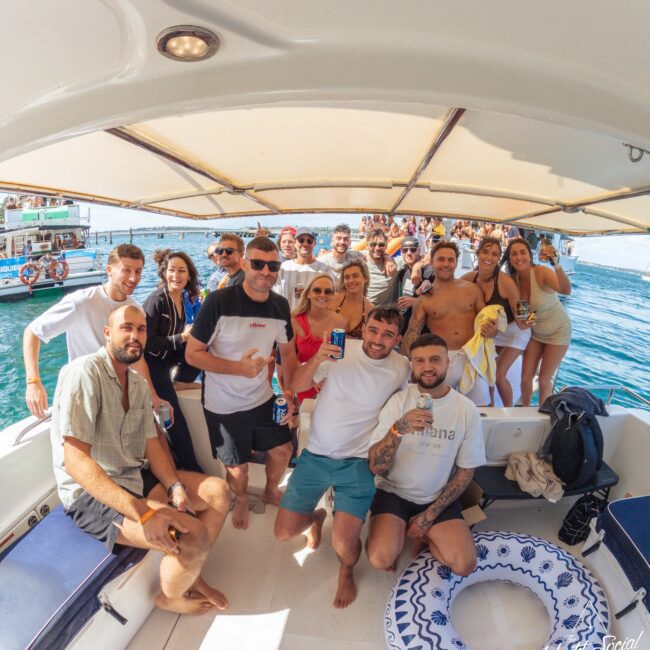 A group of people pose and smile on a boat under a canopy, holding drinks with the ocean and other boats in the background, enjoying a sunny day. An inflatable float is on the deck in front.