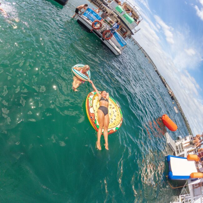 Two women relax on colorful pool floats in a large lake near a docked party boat. One woman lays on a pizza-shaped float, holding hands with the other on a donut float. Other boats and people are visible in the background.