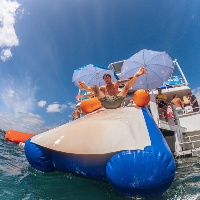 A man in swimwear slides down an inflatable water slide from a boat into the ocean, arms outstretched, with other people relaxing and blue umbrellas on the boat under a partly cloudy sky.