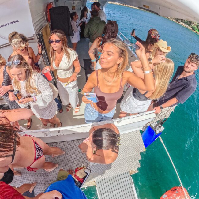 A group of young adults enjoy a lively boat party under the sun, some dancing and others socializing, with clear blue water and green shoreline visible in the background.