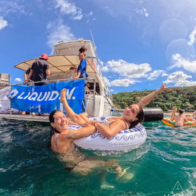 Two women smiling and posing on an inflatable ring in the water near a boat, with people on the boat and in the water. The sky is blue with clouds and the scene is lively and cheerful.
