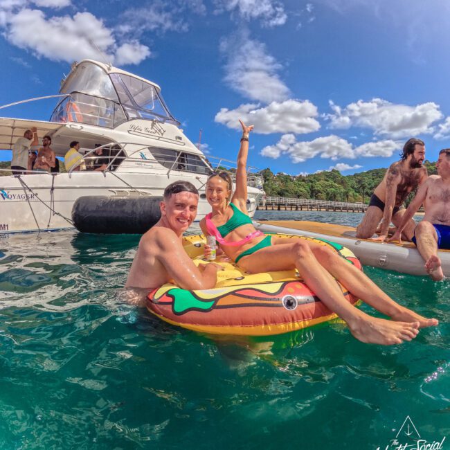 Two people smile and pose on a colorful inflatable in the water near a boat, with others relaxing nearby. The sky is blue with fluffy clouds, and there are trees on the shore in the background.
