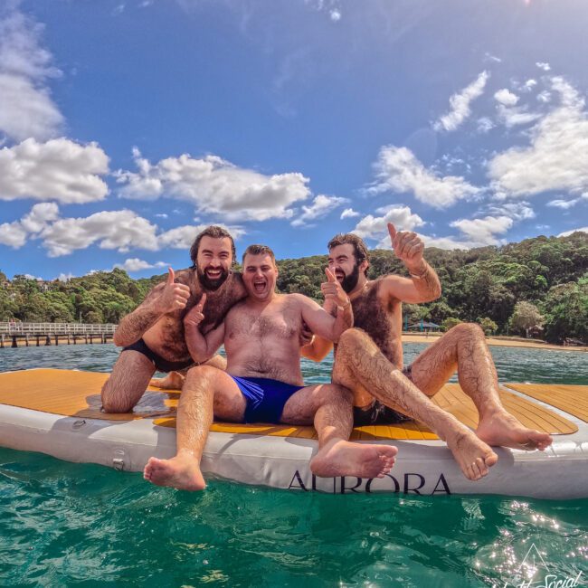Three smiling men in swim trunks sit on a floating dock in clear blue water, giving thumbs up. Behind them are green trees, a pier, and a bright, partly cloudy sky.