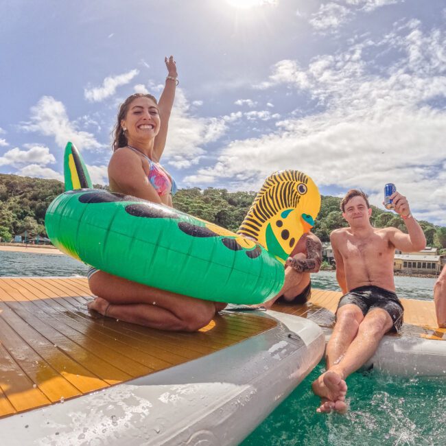 A woman in a green inflatable float and a man holding a can sit on a dock by the water, smiling and enjoying a sunny day with blue skies and scattered clouds.