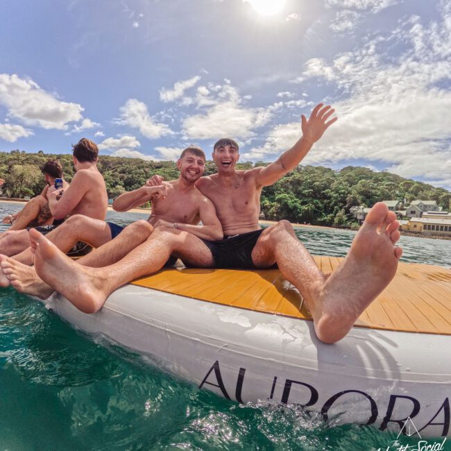 Two smiling men sit on a paddleboard in the water, waving at the camera, with two more people behind them. The sun shines above, trees and buildings are visible in the background. The word "AURORA" is on the board.