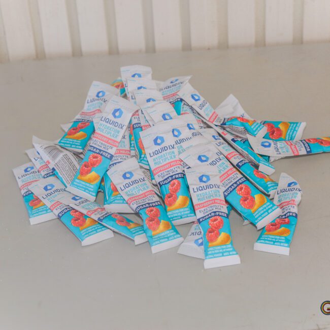A pile of Liquid IV Hydration Multiplier single-serve packets with strawberry flavor lies scattered on a light gray table. The packets are mostly white with blue, red, and orange accents.