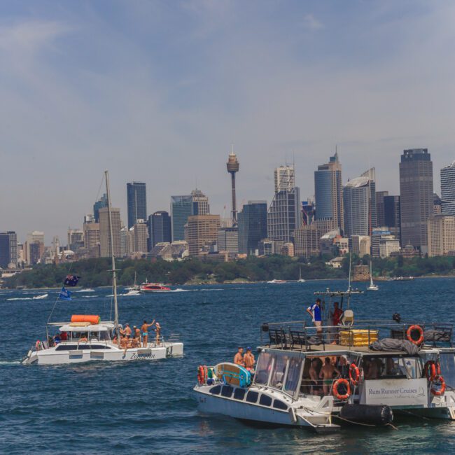 Boats with people enjoying on the water, with a city skyline and tall buildings, including a distinctive tall tower, visible in the background under a clear sky.
