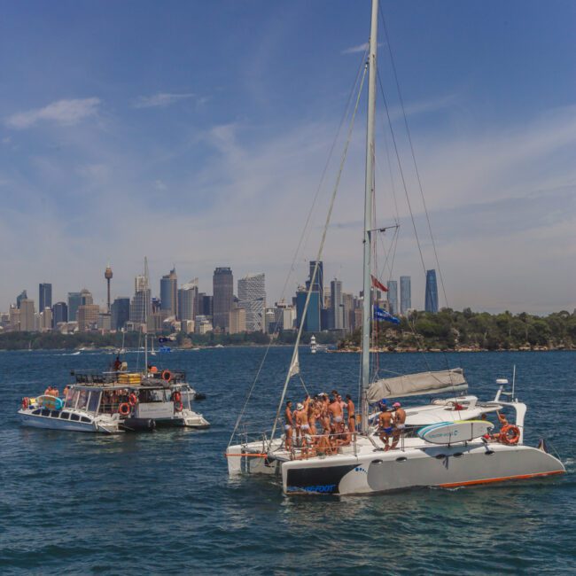 A group of people relax on a white catamaran and a nearby boat on blue water, with a city skyline and tall buildings in the background under a clear sky.