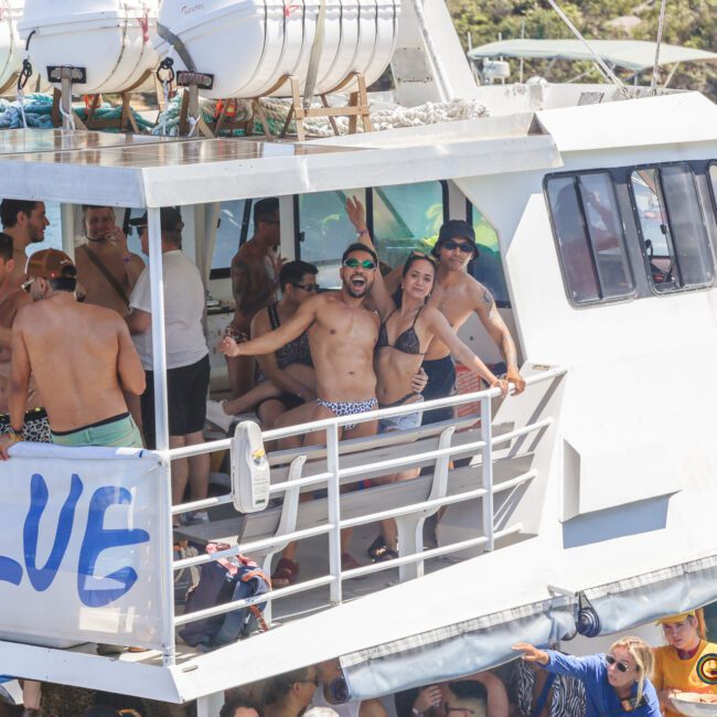 A group of young people in swimwear enjoy a lively party on a white boat named "BLUE" under sunny weather, with some standing on the deck and others inside, with water and greenery in the background.