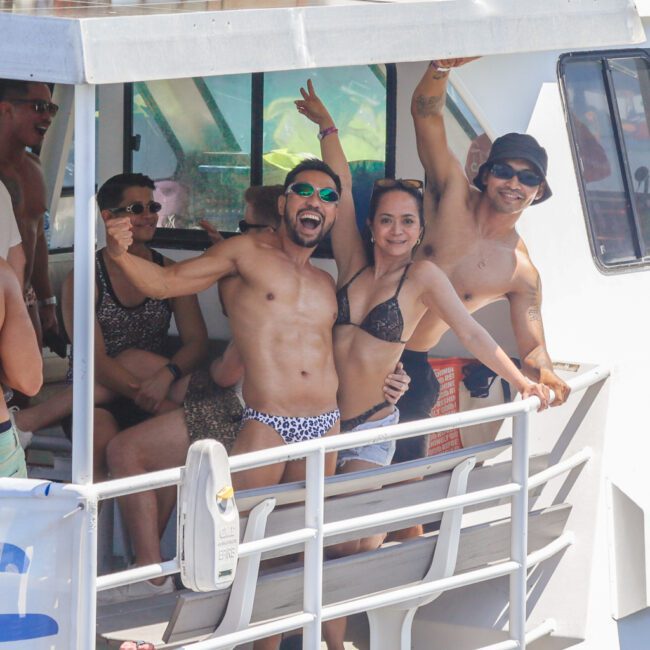 A group of people in swimsuits smile and pose energetically on the deck of a boat, enjoying a sunny day. Some wave at the camera; the atmosphere is lively and festive. The logo "GAYM" is visible in the bottom right corner.