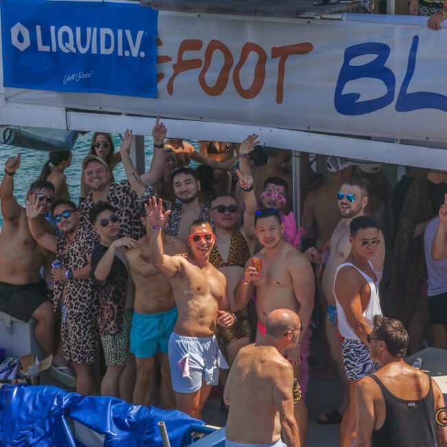 A group of people in swimwear and animal-print outfits are gathered on a boat, smiling and waving at the camera. A banner above them reads "FOOT BLUE." The boat is on the water under sunny weather.
