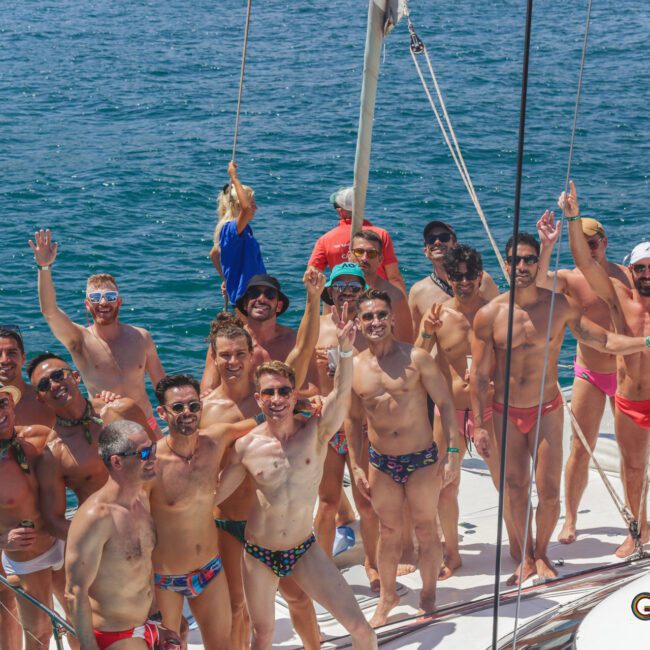 A group of men in swimwear smile and wave while standing on a sailboat in clear blue water under a sunny sky. Some wear sunglasses and hats. The atmosphere is festive and joyful.