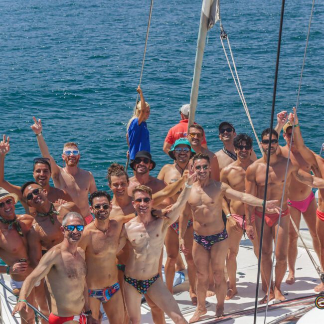 A group of men in swim trunks pose cheerfully on the deck of a sailboat in sunny weather, with clear blue water in the background. Most are smiling, raising their arms, and appearing to enjoy a fun day at sea.