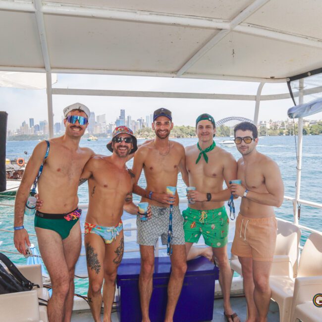 Five men in colorful swimwear pose and smile together on a boat, holding drinks. The sunny city skyline, blue sky, and water are visible in the background, with other boats nearby.