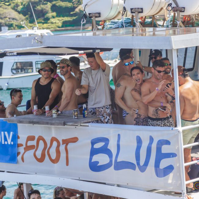 A group of men in swimwear, many wearing leopard-print shorts, party and pose on a boat decorated with a blue "LIQUID I.V." banner and a sign reading "FOOT BLUE," surrounded by water and other boats.