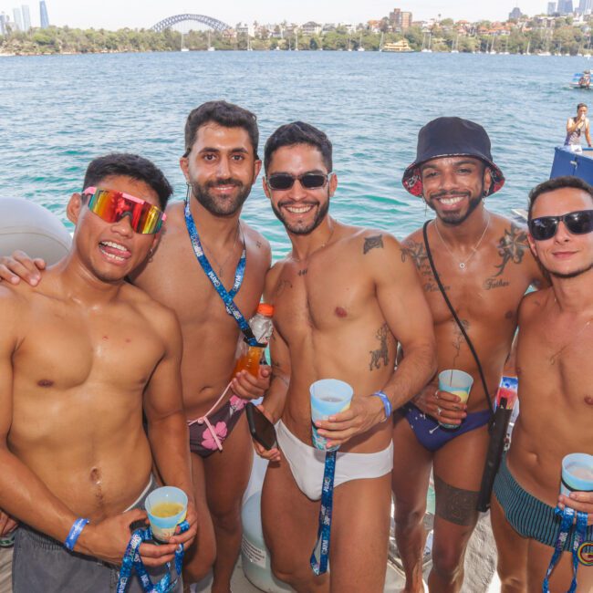 A group of six men in swimwear stand smiling together on a sunny boat party, holding drinks, with a scenic waterfront and city skyline in the background.