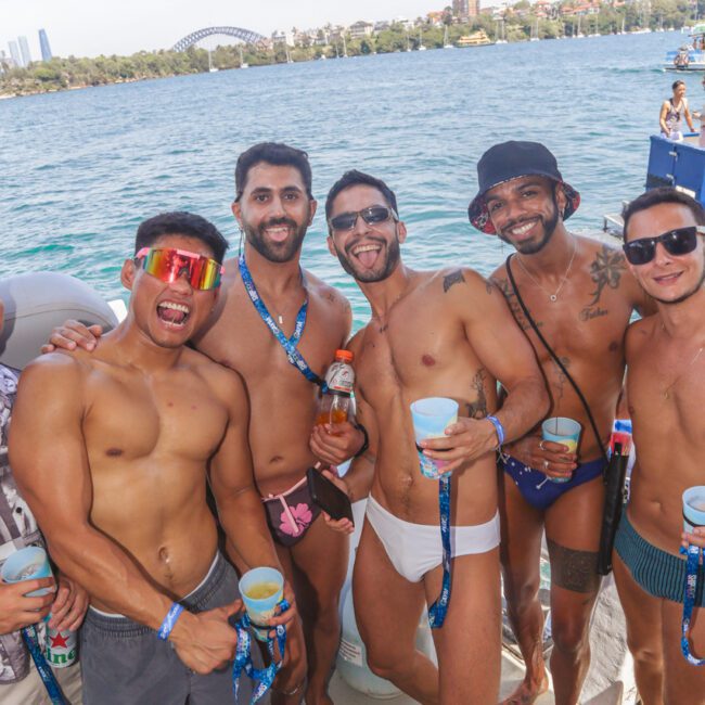 A group of five men in swimwear smile and pose together on a boat, holding drinks. Water and city buildings are visible in the background. It appears to be a sunny day and a festive, social atmosphere.