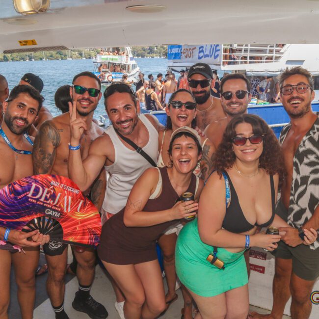 A group of people in swimsuits and summer outfits smile and pose together on a boat during a lively party, with water and other boats visible in the background.