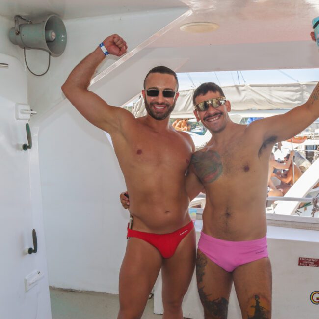 Two men in swimsuits stand on a boat, smiling and posing with their arms raised. One wears red swim briefs and sunglasses, the other wears pink briefs and holds a drink. Other people and the boat deck are visible in the background.