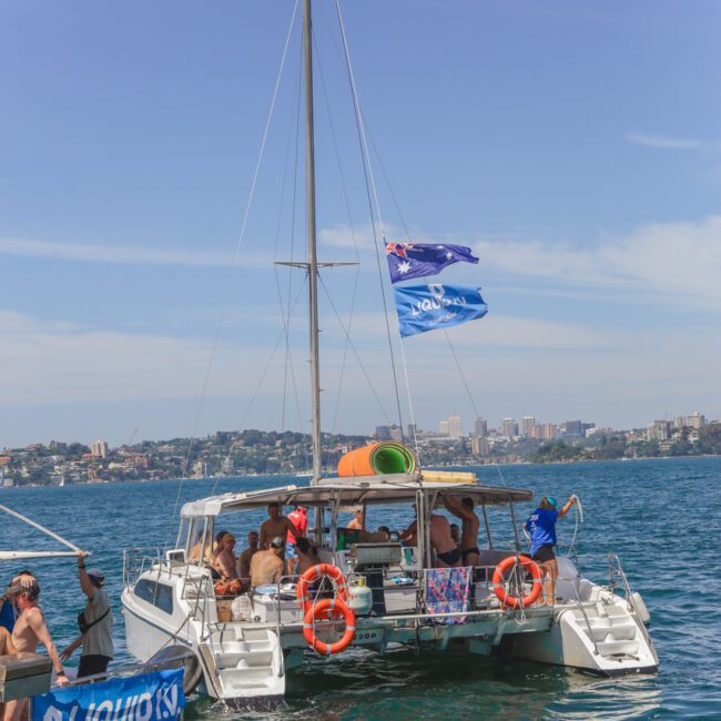 A catamaran with blue flags and orange life rings floats on the water, crowded with people in swimwear. Another boat with a “LIQUID” banner is nearby. City buildings and greenery are visible on the distant shore.