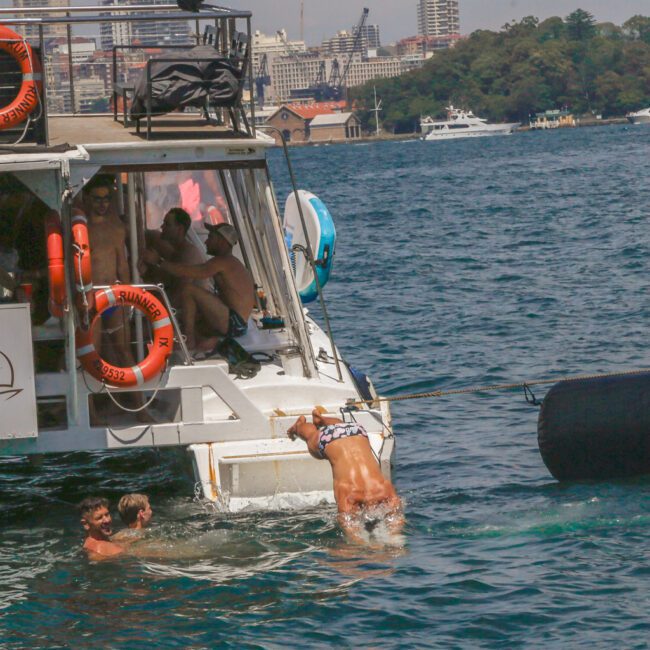 A person in swimwear climbs onto a boat from the water, while two others swim nearby. Several people relax on the boat deck. City buildings and another boat are visible in the background across the water.
