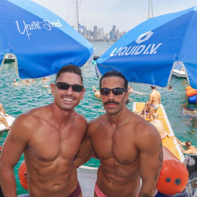 Two shirtless men with sunglasses stand smiling under blue umbrellas on a boat, with people lounging and swimming in the water nearby. City skyline and sailboats are visible in the background.