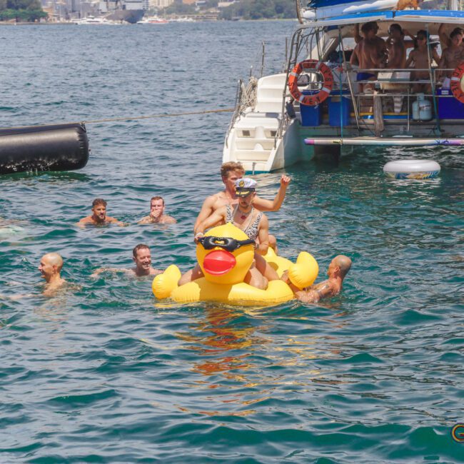 A group of people swim and play in the water near a boat. One person rides an inflatable yellow duck, wearing a crown and sash. The scene is lively and festive with a city skyline in the background.