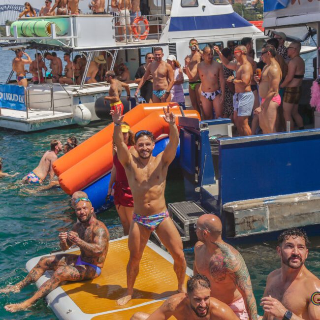 A lively group of people in colorful swimsuits enjoy a boat party on the water, with some swimming and others dancing or socializing. The Sydney Harbour Bridge is visible in the background.