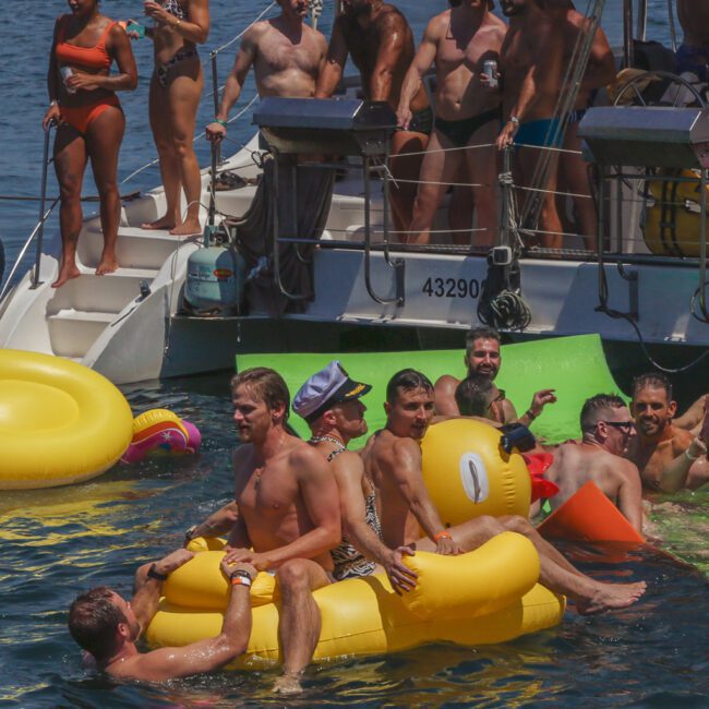 A group of people enjoy a sunny day on a boat and in the water, some lounging on inflatable floats, including a yellow duck, while others stand on the boat or swim nearby, all appearing relaxed and festive.