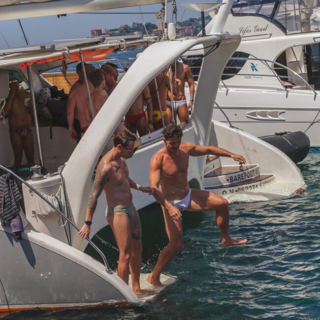 Two men in swimwear step off the back of a boat into the water, while several other people in swimwear enjoy the sunny day on the boat; nearby, another yacht is visible on the water.