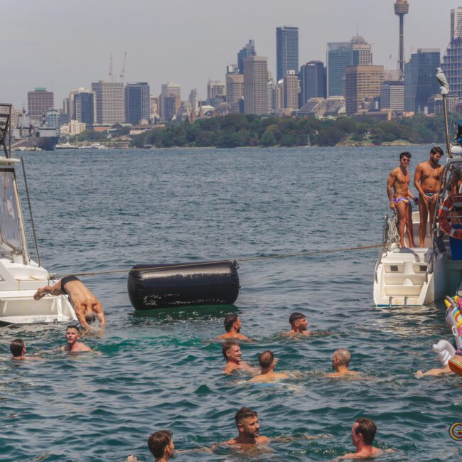 People swim and relax between two boats on the water with a city skyline in the background; some are on the boats, others are in the water, and a few are lounging on inflatables.