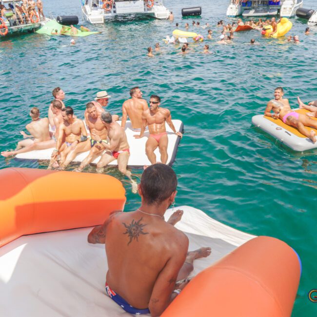 A group of people relax on floating rafts and inflatables in clear blue water near several boats. One person with a back tattoo sits at the top of an inflatable slide, about to go into the water.