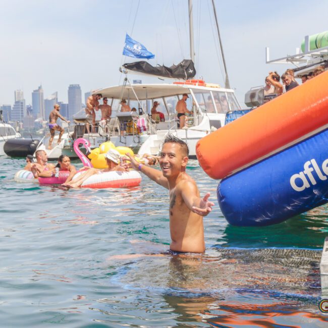 A smiling person stands waist-deep in water near colorful inflatables and boats, surrounded by people relaxing and partying on floats with a city skyline in the background. The mood is festive and lively.