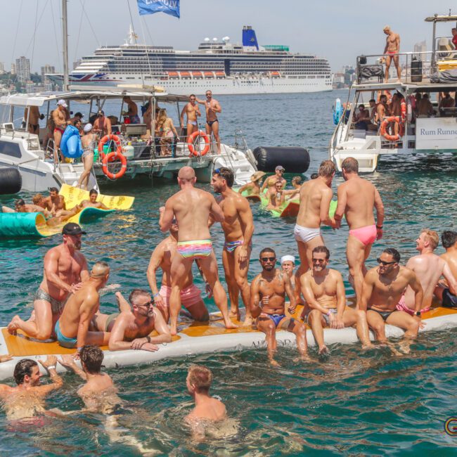 A group of men in swimwear gather on an inflatable platform and in the water, surrounded by boats during a lively, sunny boat party. A large cruise ship and city skyline are visible in the background.