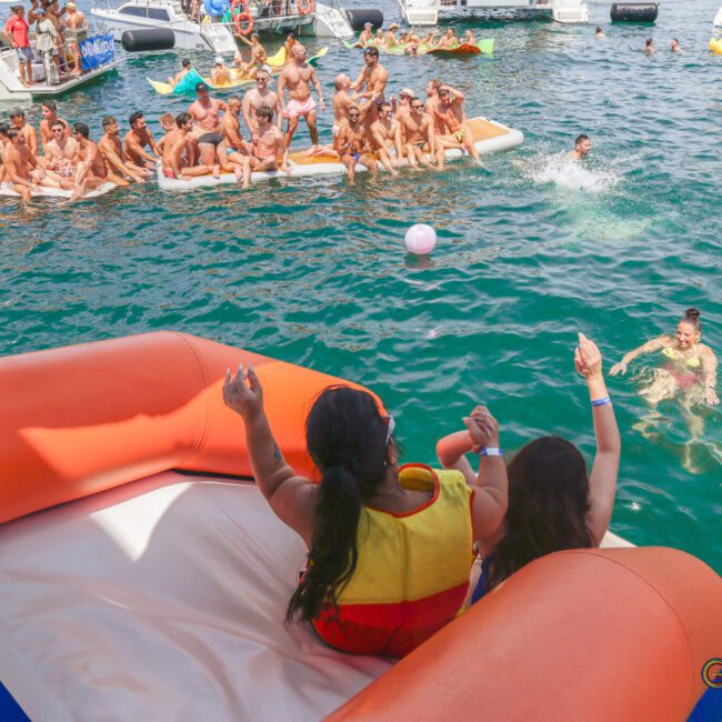 Two people on an orange inflatable slide are about to splash into a crowded, lively lake surrounded by people on floating rafts and boats under the sun.