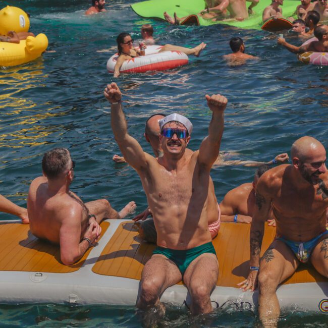 Men in swimsuits relax and celebrate on inflatable rafts and floaties in a crowded, sunny pool party. One man in the center raises his arms in excitement. The atmosphere is lively and festive.