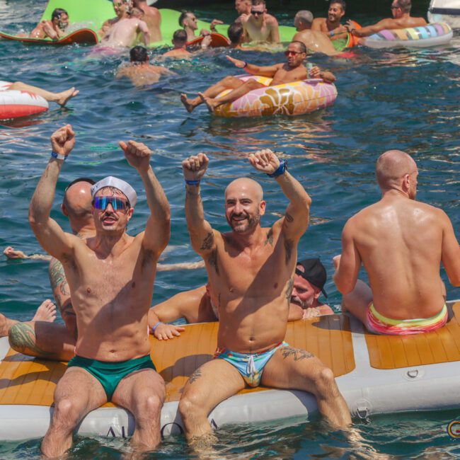 A group of people in swimsuits enjoy a pool party, with two men in the foreground raising their arms and smiling while sitting on an inflatable dock. Others float on pool rafts and swim in the water nearby.