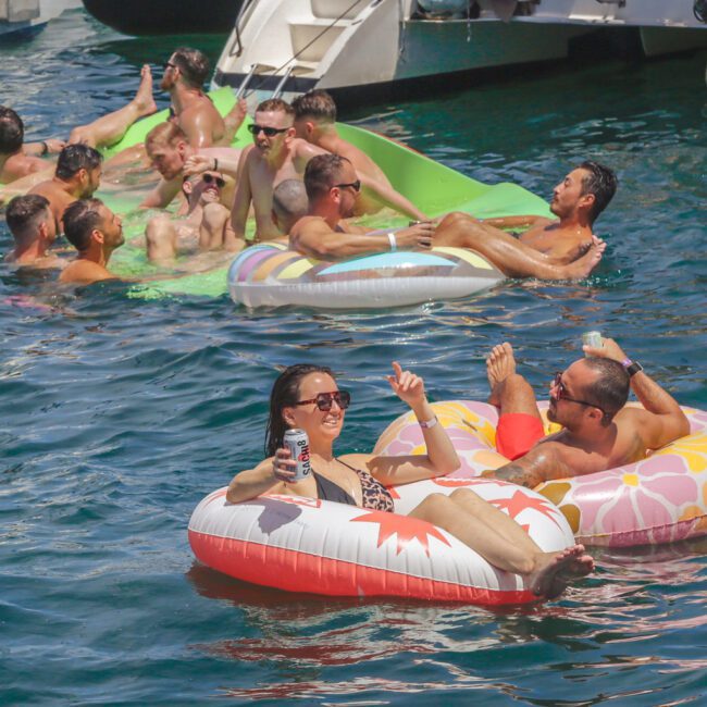 A group of people relax and socialize on colorful inflatable floats in the water near a boat. Some are holding drinks and wearing swimsuits, enjoying a sunny day. A logo reading "GAYM" is visible in the bottom right corner.