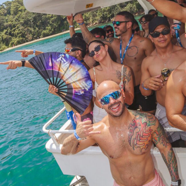 A group of people in swimwear enjoy a boat party on clear blue water, smiling and posing for the camera. One person in front gives a thumbs up; others hold drinks and a colorful fan. Lush green trees are visible in the background.