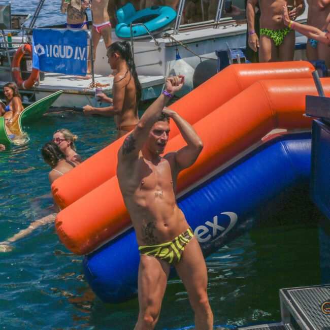 A man in a yellow patterned swimsuit dances by an inflatable slide in the water, surrounded by people on floats and boats at a lively, sunny pool party.