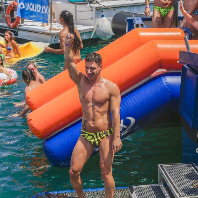 A man in a yellow patterned swimsuit stands on a boat dock, raising his arm triumphantly. Behind him, people in swimsuits relax on inflatables and boats, enjoying a sunny day on the water.
