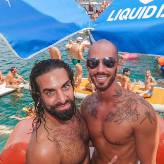 Two men pose shirtless and smiling at an outdoor pool or boat party, surrounded by people in swimwear. Blue umbrellas and city buildings are visible in the background on a sunny day.