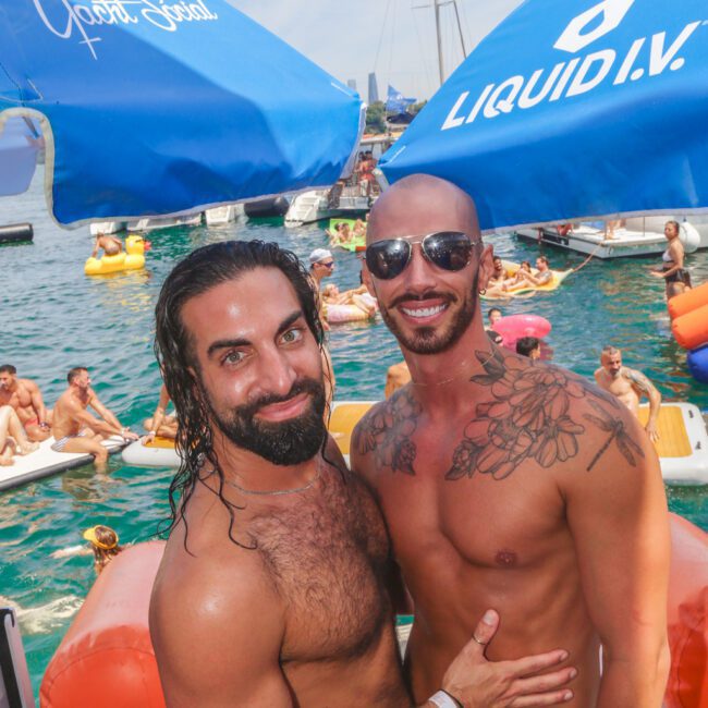 Two shirtless men smile and pose together on a boat at a lively pool party, with people swimming and relaxing on floats in the water behind them. Large blue umbrellas and yachts are visible in the background.