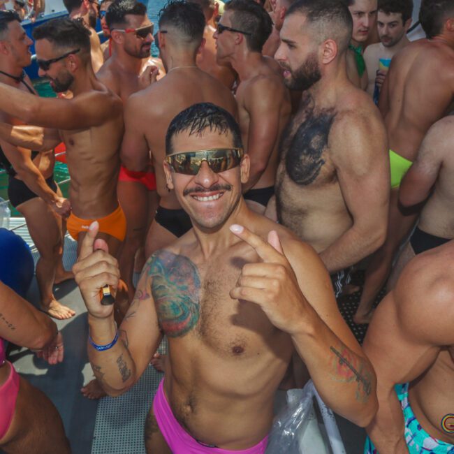 A group of shirtless men in swimwear enjoy a crowded outdoor party. One man in pink swim briefs and sunglasses smiles and points at the camera; others are chatting and dancing. The scene is lively and festive.