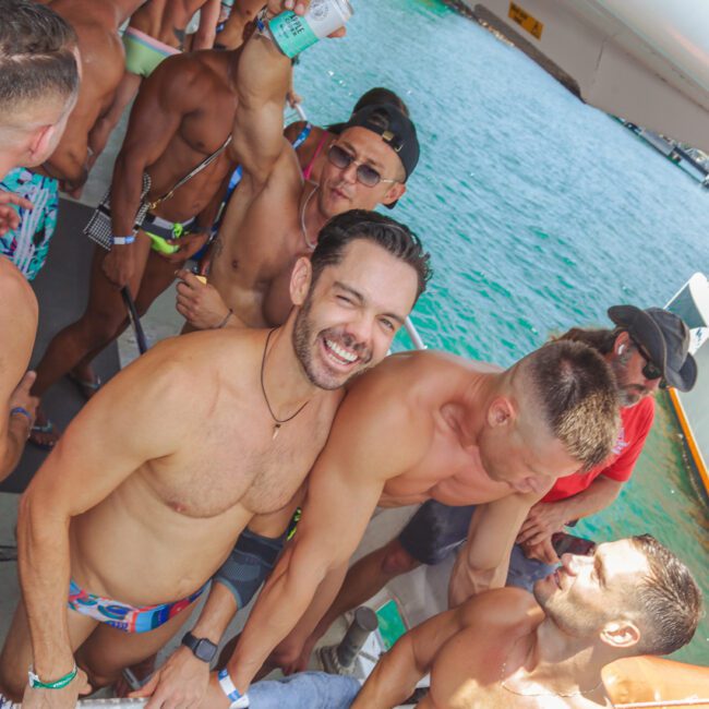 A group of shirtless men in swimwear smile and socialize on a boat by the water; one man holds up a drink, and others are gathered closely, enjoying a sunny day. The "GAYM" logo is visible in the corner.