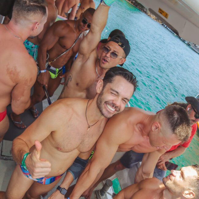 A group of shirtless men in swimwear smile and pose on a boat over turquoise water, with one man giving a thumbs up and another raising his drink in celebration.