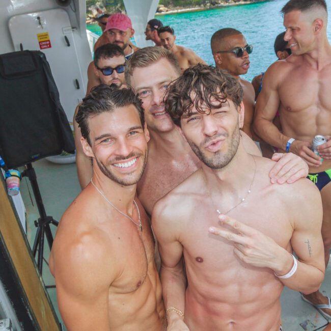A group of shirtless men pose and smile for a photo on a boat during a sunny day, with blue water and trees visible in the background. One man in front flashes a peace sign. The atmosphere appears festive and lively.